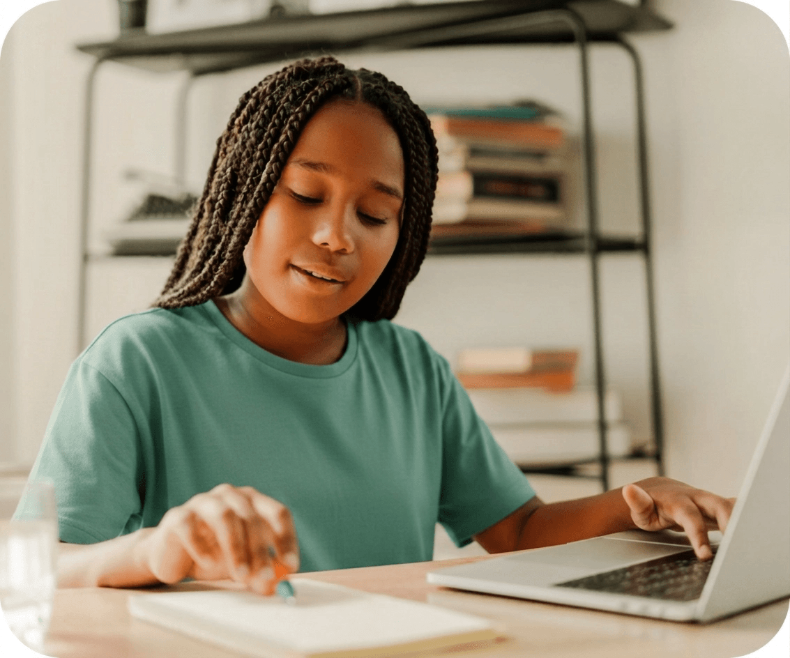 A student writes in a notebook while studying at a laptop, with books on a shelf behind her